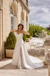 A woman in a strapless white 88515/MADISON wedding dress with lace detailing poses outside a building with arched windows, near potted plants and white metal patio furniture.