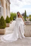 A woman in a strapless white 88515/MADISON wedding dress with lace detailing poses outside a building with arched windows, near potted plants and white metal patio furniture.