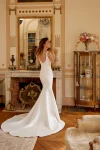 A woman in a fitted, sleeveless white gown—88516/MARLEY by Justin Alexander—stands in an elegant living room with wooden floors, large windows, and indoor plants.