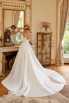 A woman in a fitted, sleeveless white gown—88516/MARLEY by Justin Alexander—stands in an elegant living room with wooden floors, large windows, and indoor plants.
