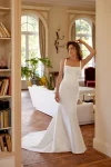 A woman in a fitted, sleeveless white gown—88516/MARLEY by Justin Alexander—stands in an elegant living room with wooden floors, large windows, and indoor plants.