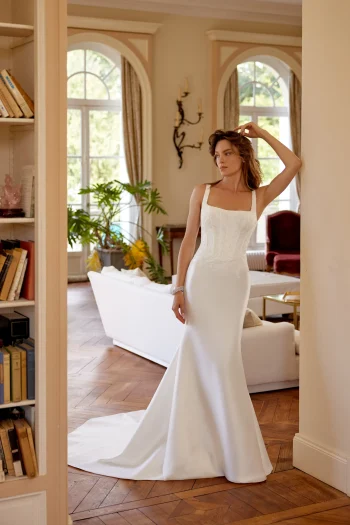 A woman in a fitted, sleeveless white gown—88516/MARLEY by Justin Alexander—stands in an elegant living room with wooden floors, large windows, and indoor plants.