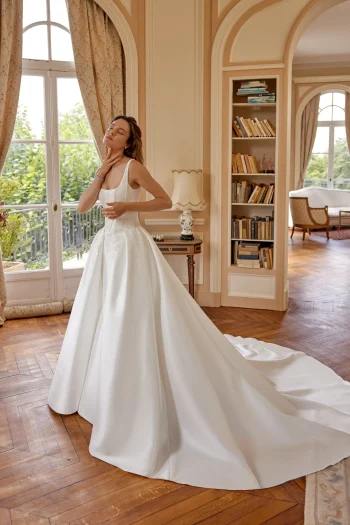 A woman in a white 88516/MARLEY by Justin Alexander wedding gown stands in a sunlit room with large windows, wood floors, and bookshelves, appearing serene and contemplative—a vision of grace reminiscent of the timeless elegance embodied by MARLEY.