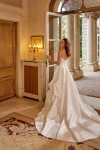 Wearing the 88519/MARA by Justin Alexander, a woman stands indoors near an open door, adjusting her hair in a strapless white wedding gown with a garden visible outside.