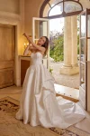 Wearing the 88519/MARA by Justin Alexander, a woman stands indoors near an open door, adjusting her hair in a strapless white wedding gown with a garden visible outside.