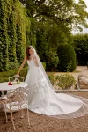 Wearing the 88519/MARA by Justin Alexander, a woman stands indoors near an open door, adjusting her hair in a strapless white wedding gown with a garden visible outside.