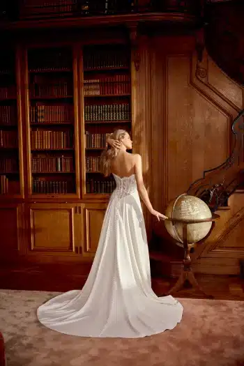 A woman in a strapless white gown stands with her back to the camera, holding a globe in a wood-paneled library lined with bookshelves.