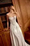 Woman in a strapless white lace and satin gown stands by wooden bookshelves, looking to the side.