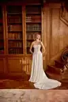Woman in a strapless white lace and satin gown stands by wooden bookshelves, looking to the side.