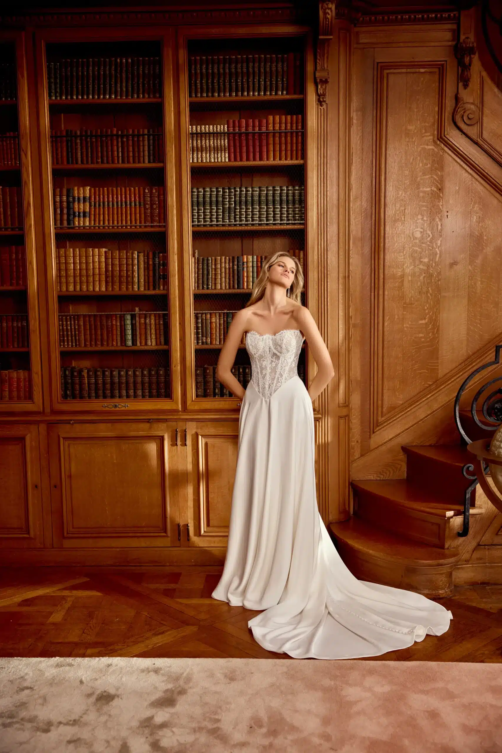 Woman in a strapless white wedding dress stands in front of a wooden bookcase filled with books, next to a staircase, in a room with wood paneling. Woman in a strapless white wedding dress stands in front of a wooden bookcase filled with books, next to a staircase, in a room with wood paneling.