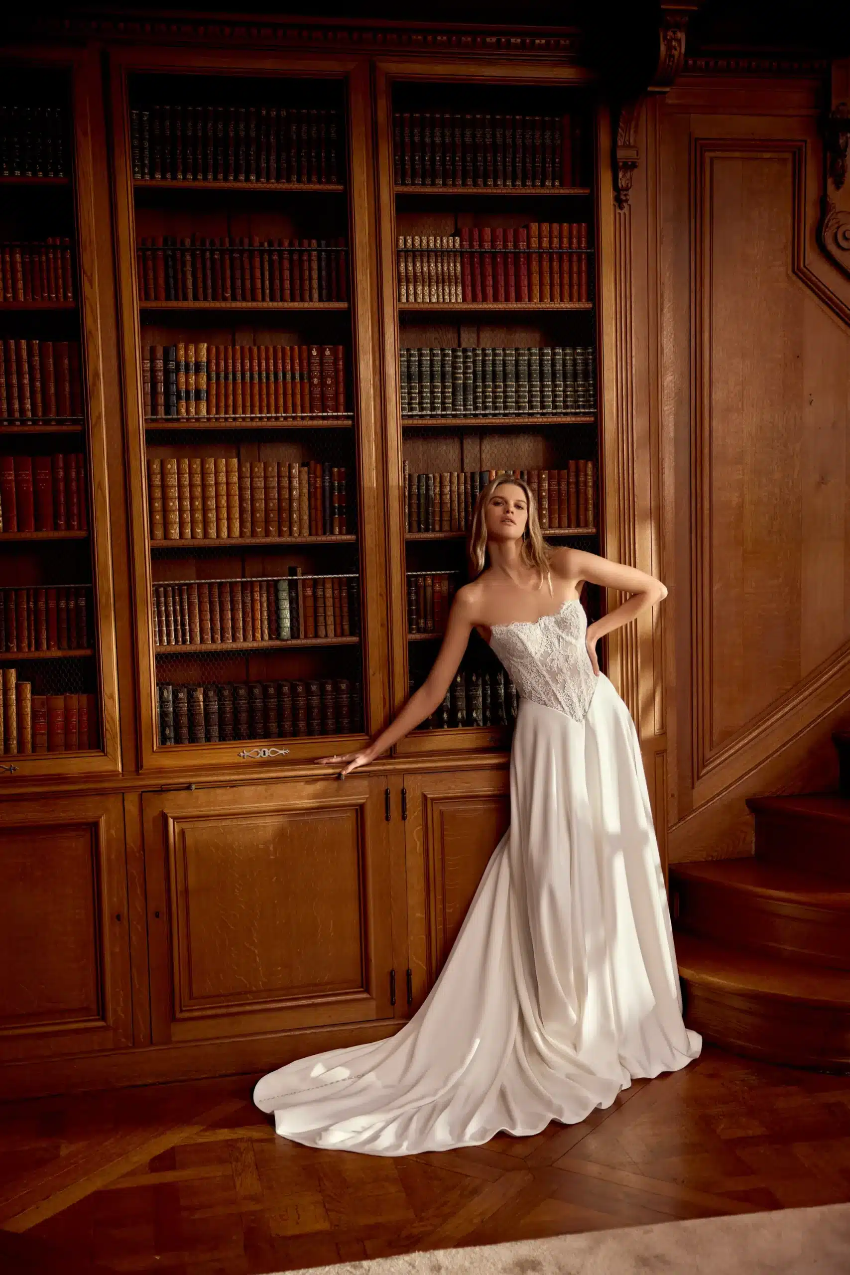 Woman in a strapless white wedding gown stands in front of a tall wooden bookcase filled with books in a wood-paneled room. Woman in a strapless white wedding gown stands in front of a tall wooden bookcase filled with books in a wood-paneled room.