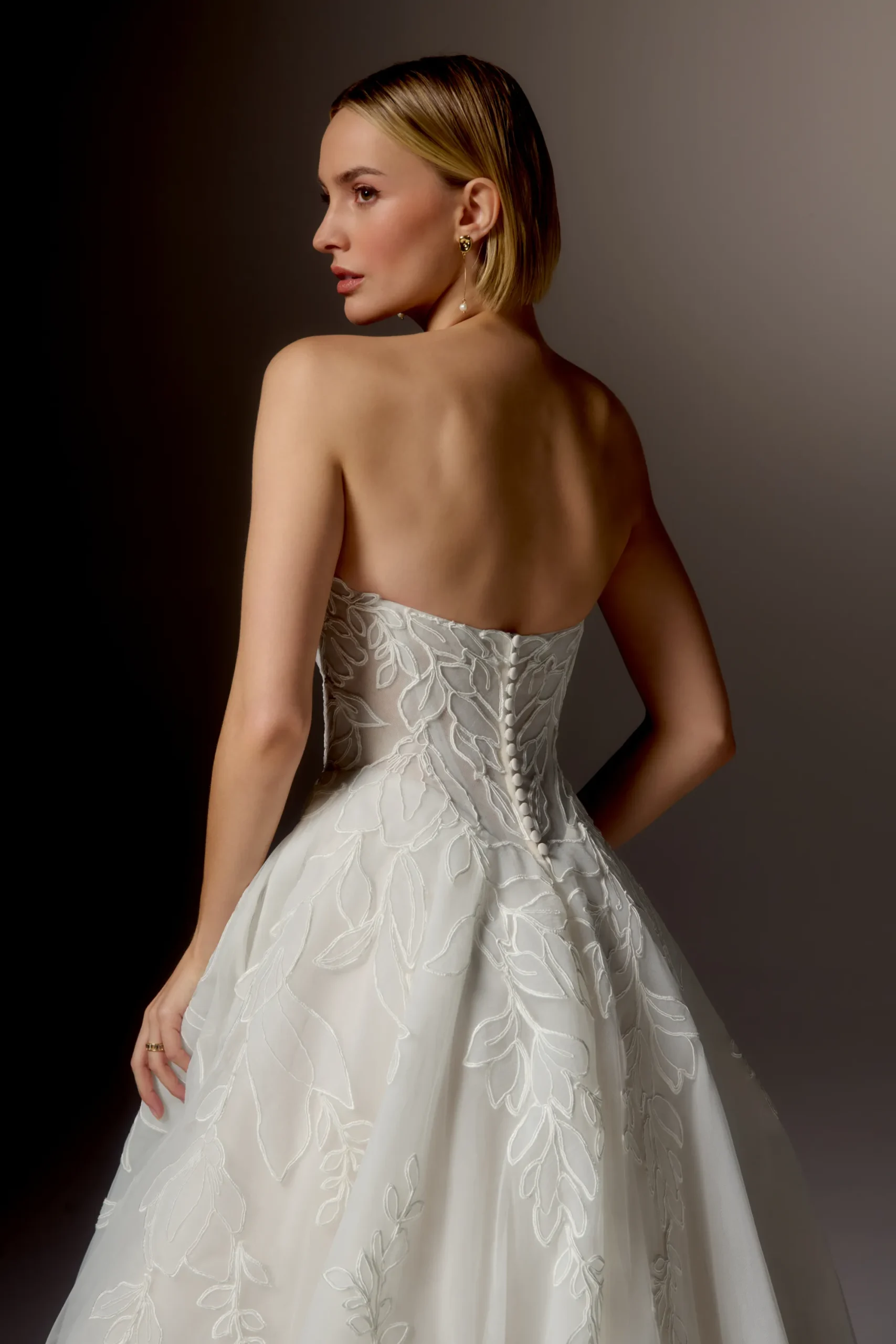 A woman in a strapless white 99405/RAVENNA by Justin Alexander wedding gown, adorned with elegant leaf patterns, gazes over her shoulder against a neutral background. This striking style showcases timeless beauty and sophistication. A woman in a strapless white 99405/RAVENNA by Justin Alexander wedding gown, adorned with elegant leaf patterns, gazes over her shoulder against a neutral background. This striking style showcases timeless beauty and sophistication.