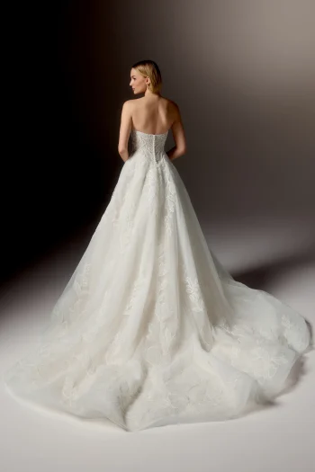 A woman stands facing away, wearing the sophisticated 99405/RAVENNA by Justin Alexander, a strapless white wedding gown featuring a long, flowing train and delicate floral patterns.