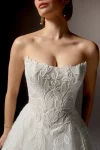 A woman stands gracefully against a neutral background, wearing the 99405/RAVENNA strapless white wedding gown by Justin Alexander, featuring delicate floral patterns and paired with a long sheer veil.