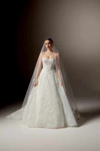 A woman stands gracefully against a neutral background, wearing the 99405/RAVENNA strapless white wedding gown by Justin Alexander, featuring delicate floral patterns and paired with a long sheer veil.