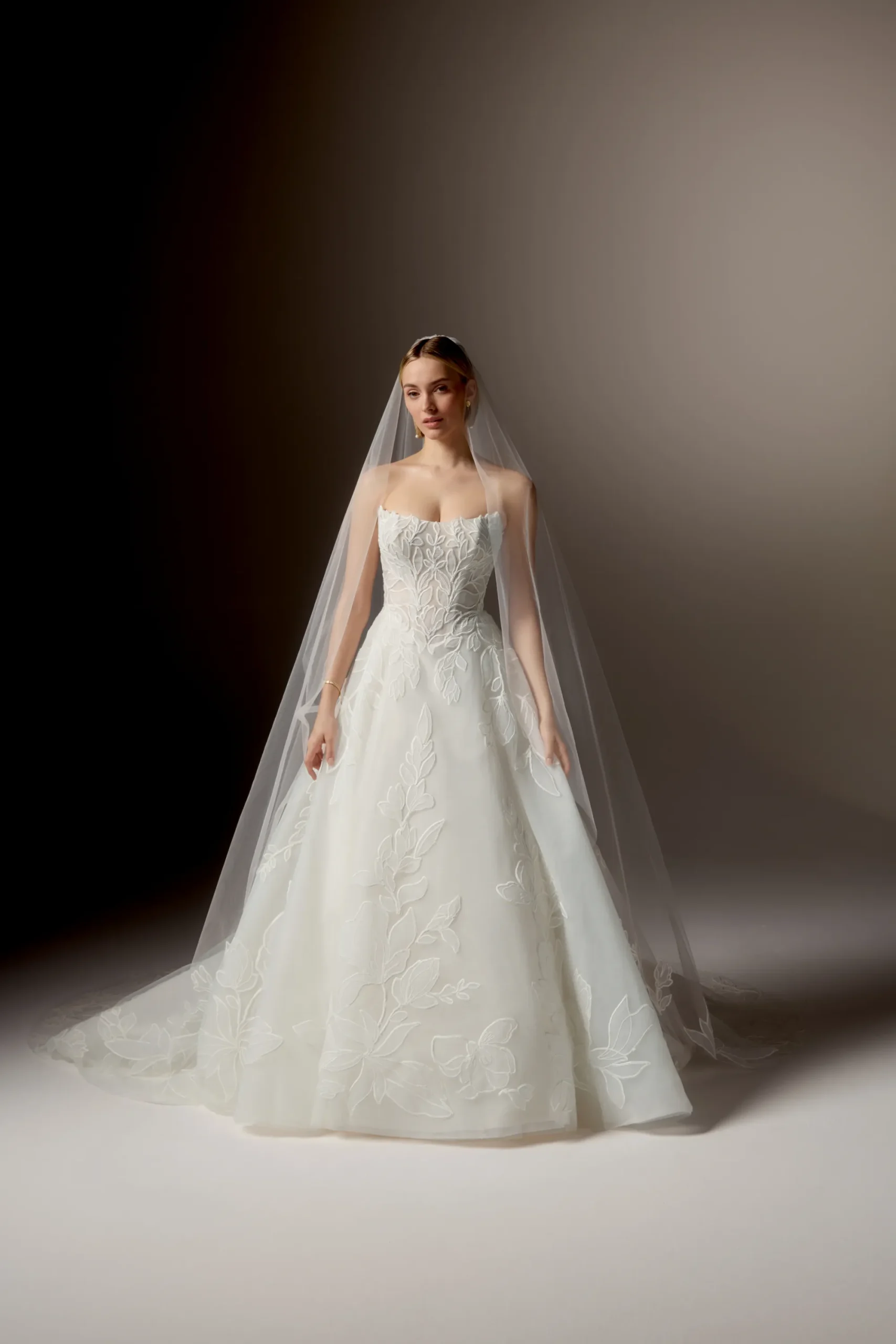 A woman stands gracefully against a neutral background, wearing the 99405/RAVENNA strapless white wedding gown by Justin Alexander, featuring delicate floral patterns and paired with a long sheer veil. A woman stands gracefully against a neutral background, wearing the 99405/RAVENNA strapless white wedding gown by Justin Alexander, featuring delicate floral patterns and paired with a long sheer veil.