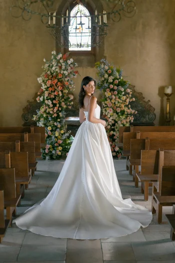 A woman in a white wedding dress stands in a church aisle, facing backward, with flower arrangements and stained glass in the background.
