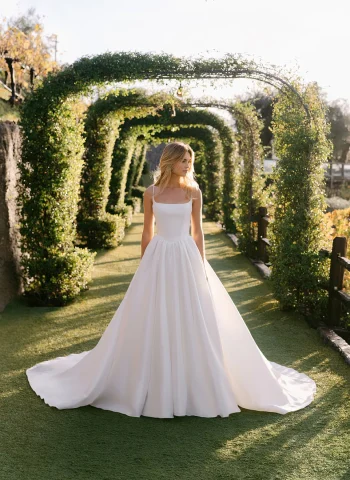 Wearing the A1402 by Allure, a woman stands on a garden path beneath green archways, looking to the side in natural sunlight.
