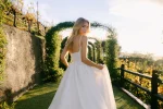 Wearing the A1402 by Allure, a woman stands on a garden path beneath green archways, looking to the side in natural sunlight.