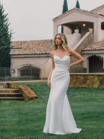 A woman exudes allure in the A1488 by Allure, a strapless white dress, as she stands on grass before a stone building with arched windows and a tiled roof.