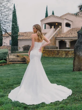 A woman wearing the A1488 by Allure, a strapless white wedding dress, stands on grass facing away, with a large stone house and trees in the background.