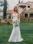 A woman exudes allure in the A1488 by Allure, a strapless white dress, as she stands on grass before a stone building with arched windows and a tiled roof.