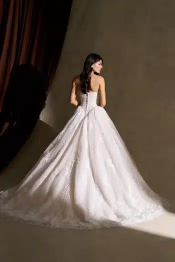 A woman wearing the C845 by Allure, a strapless lace ballgown wedding dress, stands with her back to the camera in front of a beige wall and brown curtain.