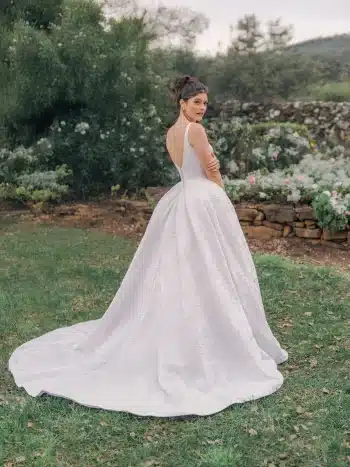 A woman in the R3987 by Allure, a white, backless wedding gown, stands outdoors on grass with her back to the camera, surrounded by flowers and greenery.
