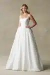 A woman stands against a neutral textured background, wearing the R3987 bridal gown by Allure—a white, sleeveless dress featuring a square neckline and a full skirt.