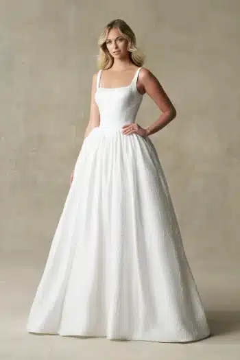 A woman stands against a neutral textured background, wearing the R3987 bridal gown by Allure—a white, sleeveless dress featuring a square neckline and a full skirt.