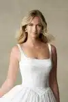 A woman stands against a neutral textured background, wearing the R3987 bridal gown by Allure—a white, sleeveless dress featuring a square neckline and a full skirt.