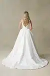 A woman stands against a neutral textured background, wearing the R3987 bridal gown by Allure—a white, sleeveless dress featuring a square neckline and a full skirt.