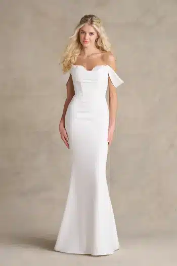 A woman stands against a neutral background wearing the R3989 by Allure, an off-the-shoulder, fitted white bridal gown featuring a subtle mermaid silhouette that exudes elegance.