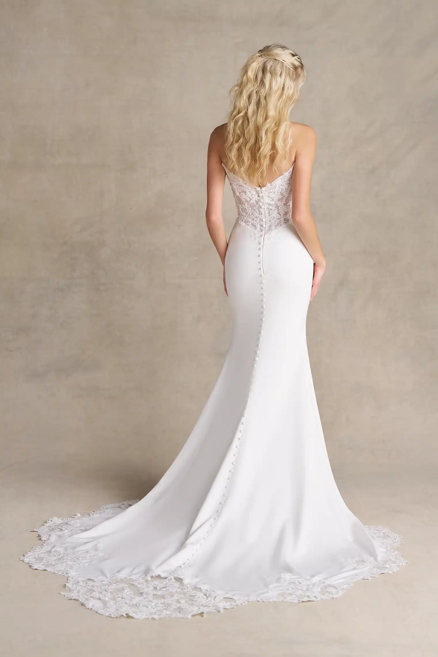 A woman stands with her back to the camera, showcasing the stunning R3989 by Allure bridal gown, featuring elegant white lace and satin, a dramatic long train, and delicate button details. A woman stands with her back to the camera, showcasing the stunning R3989 by Allure bridal gown, featuring elegant white lace and satin, a dramatic long train, and delicate button details.