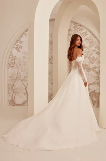 A woman stands under arched architecture, facing away from the camera, wearing the M2532 by Mikaella Bridal—an off-the-shoulder white gown with a long train.
