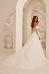A woman stands indoors against a patterned wall, wearing the M2532 by Mikaella Bridal—an off-shoulder white gown featuring lace sleeves and a thigh-high slit.