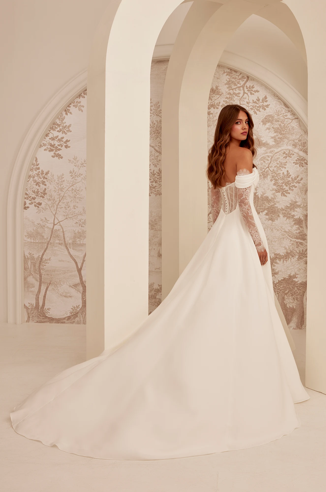 A woman stands under arched architecture, facing away from the camera, wearing the M2532 by Mikaella Bridal—an off-the-shoulder white gown with a long train. A woman stands under arched architecture, facing away from the camera, wearing the M2532 by Mikaella Bridal—an off-the-shoulder white gown with a long train.