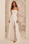 A woman stands indoors against a patterned wall, wearing the M2532 by Mikaella Bridal—an off-shoulder white gown featuring lace sleeves and a thigh-high slit.