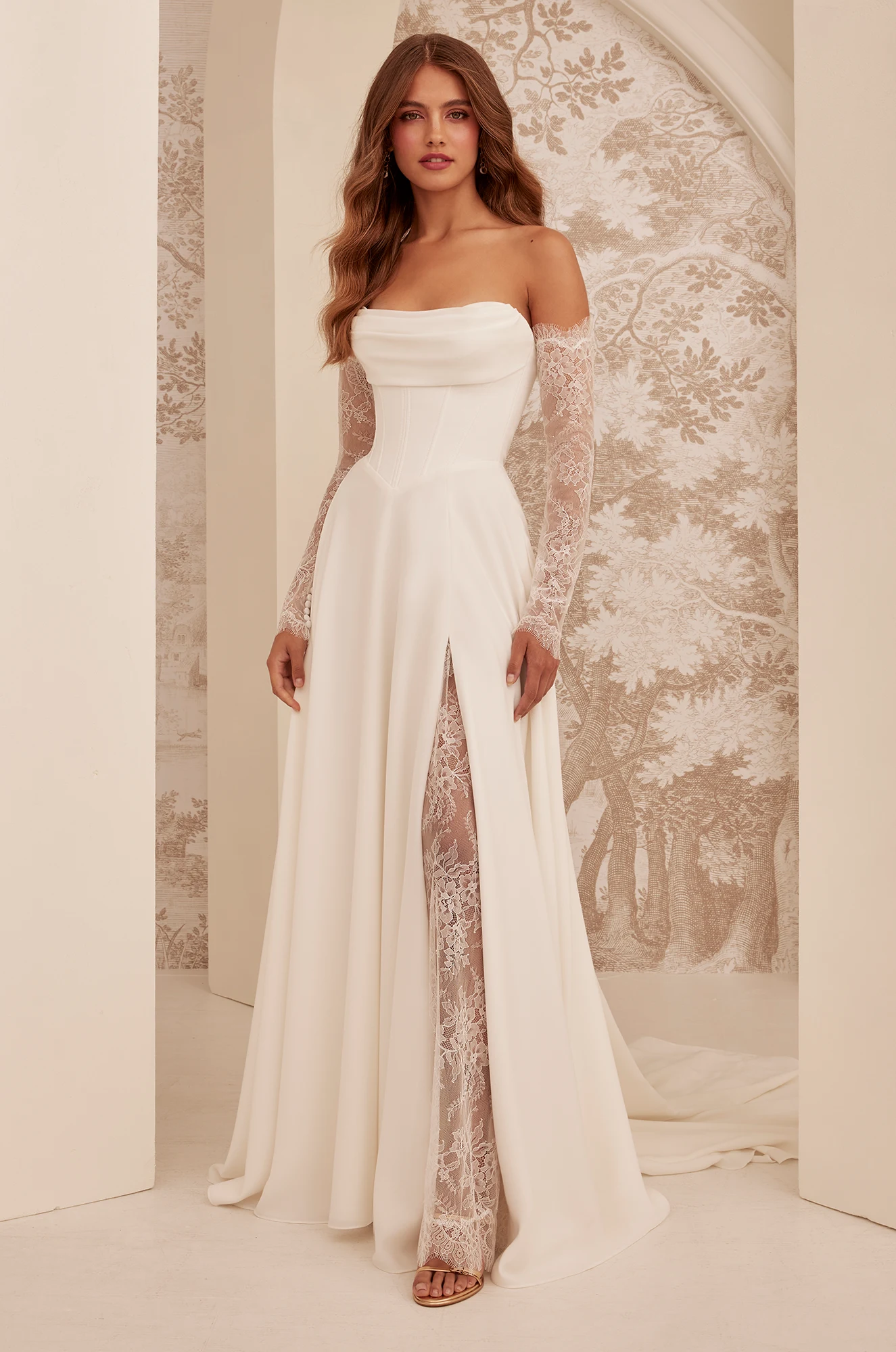 A woman wearing an off-the-shoulder white gown with lace long sleeves and a high slit stands in front of a patterned backdrop. This elegant bridal look features the M2532 by Mikaella Bridal. A woman wearing an off-the-shoulder white gown with lace long sleeves and a high slit stands in front of a patterned backdrop. This elegant bridal look features the M2532 by Mikaella Bridal.