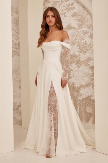 A woman stands indoors against a patterned wall, wearing the M2532 by Mikaella Bridal—an off-shoulder white gown featuring lace sleeves and a thigh-high slit.