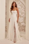 A woman stands indoors against a patterned wall, wearing the M2532 by Mikaella Bridal—an off-shoulder white gown featuring lace sleeves and a thigh-high slit.