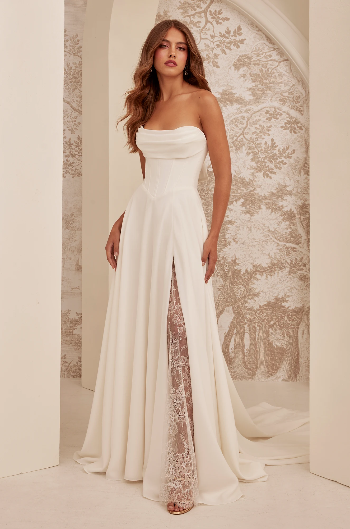 A woman stands indoors against decorative wallpaper, wearing the M2532 by Mikaella Bridal—a strapless white gown featuring a draped bodice and a lace-paneled front slit. A woman stands indoors against decorative wallpaper, wearing the M2532 by Mikaella Bridal—a strapless white gown featuring a draped bodice and a lace-paneled front slit.