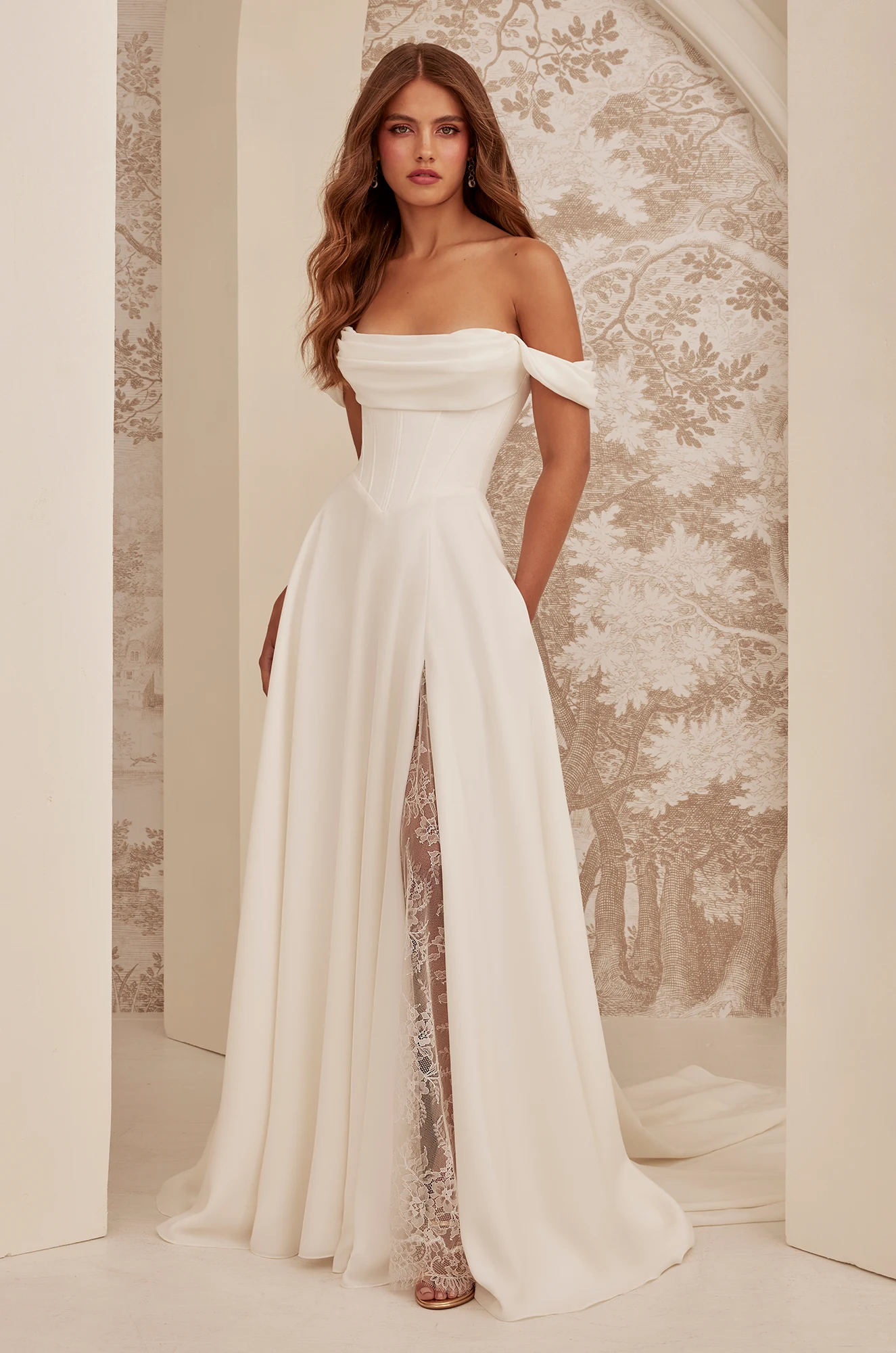 Woman wearing the M2532 by Mikaella Bridal—an off-the-shoulder white bridal gown with a corset bodice, high slit, and lace underskirt—standing before a decorative wall adorned with a leafy tree pattern. Woman wearing the M2532 by Mikaella Bridal—an off-the-shoulder white bridal gown with a corset bodice, high slit, and lace underskirt—standing before a decorative wall adorned with a leafy tree pattern.
