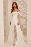A woman stands indoors against a patterned wall, wearing the M2532 by Mikaella Bridal—an off-shoulder white gown featuring lace sleeves and a thigh-high slit.