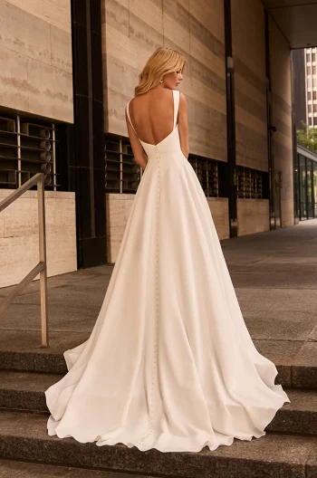 A woman stands on outdoor stone steps near a modern building, wearing the M2582 by Mikaella Bridal—a white sleeveless bridal gown with a deep V-back and buttons cascading down the train.
