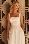 A woman stands outdoors on a city sidewalk near modern buildings, wearing the M2582 by Mikaella Bridal—a sleeveless, square-neck white bridal gown.