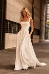 A woman stands outdoors on a city sidewalk near modern buildings, wearing the M2582 by Mikaella Bridal—a sleeveless, square-neck white bridal gown.