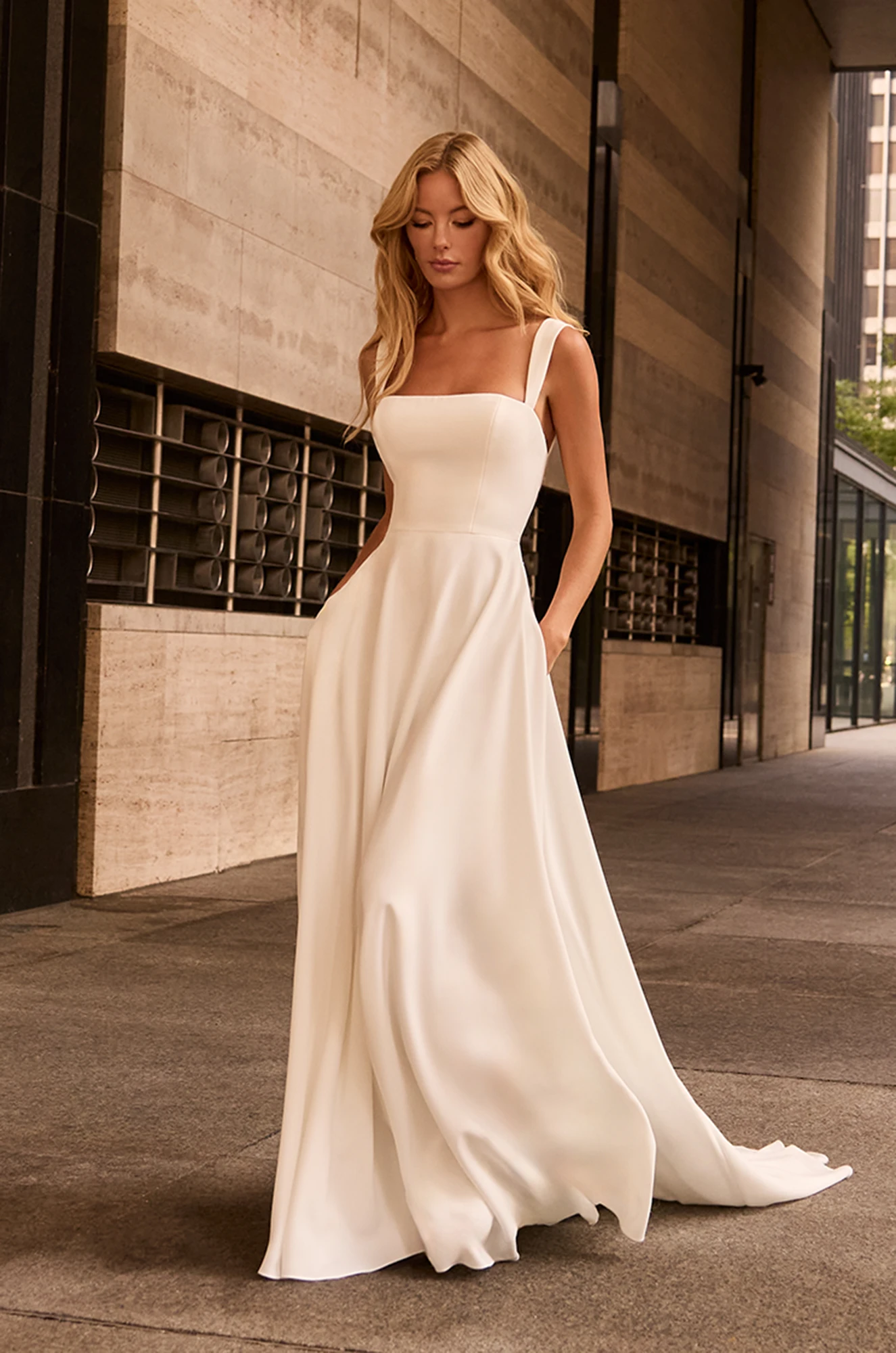 A woman stands on a sidewalk in front of a modern building with stone walls and glass windows, wearing the M2582 by Mikaella Bridal—an elegant, sleeveless, floor-length white bridal gown. A woman stands on a sidewalk in front of a modern building with stone walls and glass windows, wearing the M2582 by Mikaella Bridal—an elegant, sleeveless, floor-length white bridal gown.