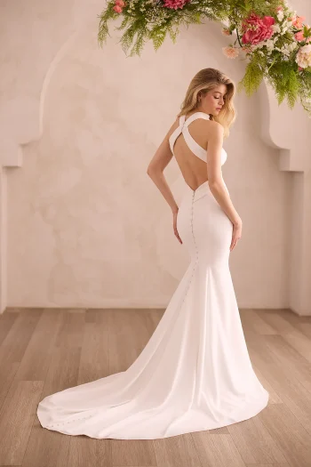 Wearing the M2604 by Mikaella Bridal, a woman stands in a fitted white gown with a long train beneath hanging flowers, facing away in a softly lit room with light-colored walls.