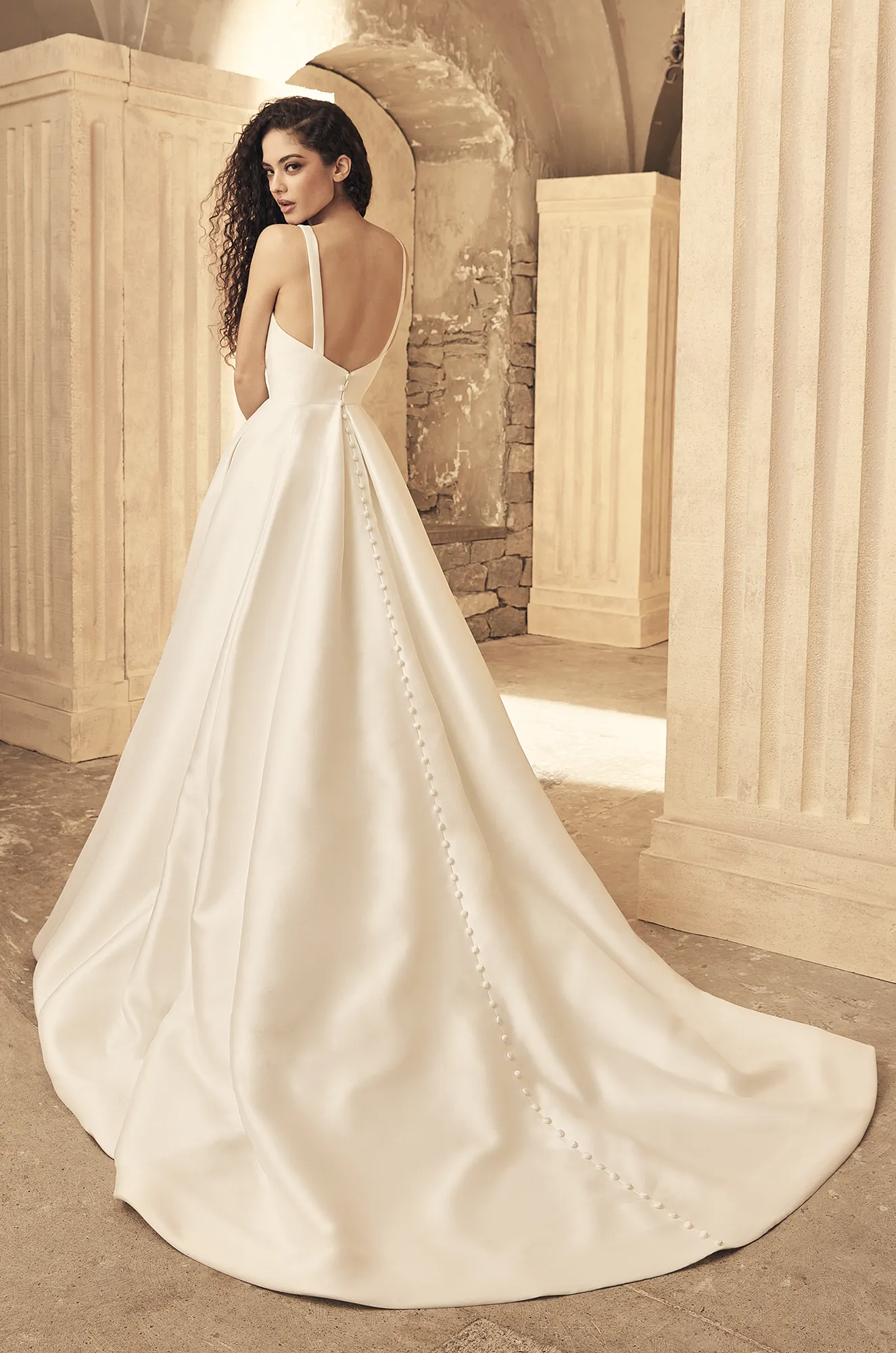A woman stands in a classical stone setting, facing away from the camera, wearing the P5150 by Paloma Blanca—a long, white satin gown featuring a low back and elegant button details. A woman stands in a classical stone setting, facing away from the camera, wearing the P5150 by Paloma Blanca—a long, white satin gown featuring a low back and elegant button details.